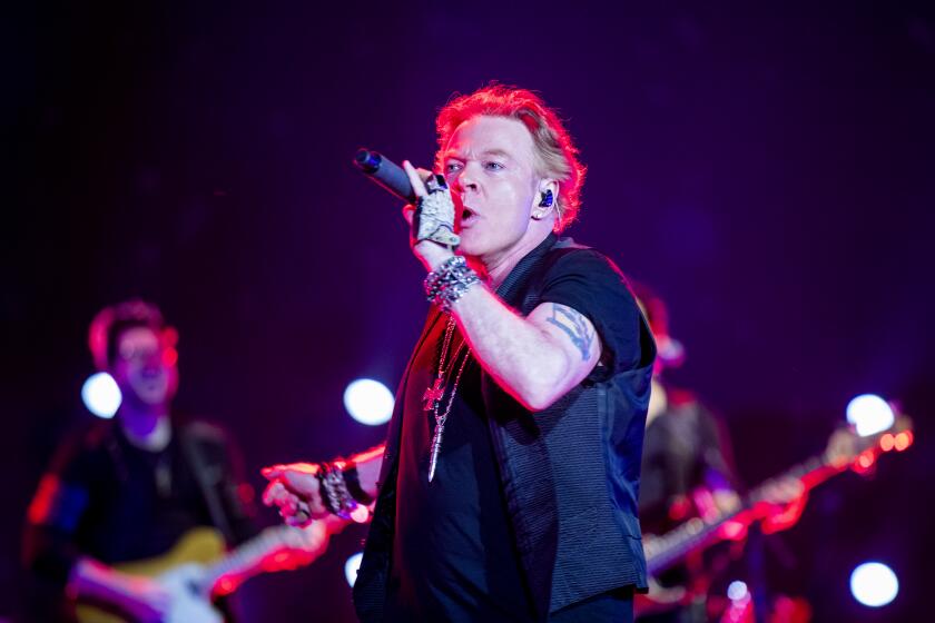 
Guns N' Roses lead singer Axl Rose in a short-sleeve black shirt holding a mic to his mouth as he performs on a stage.