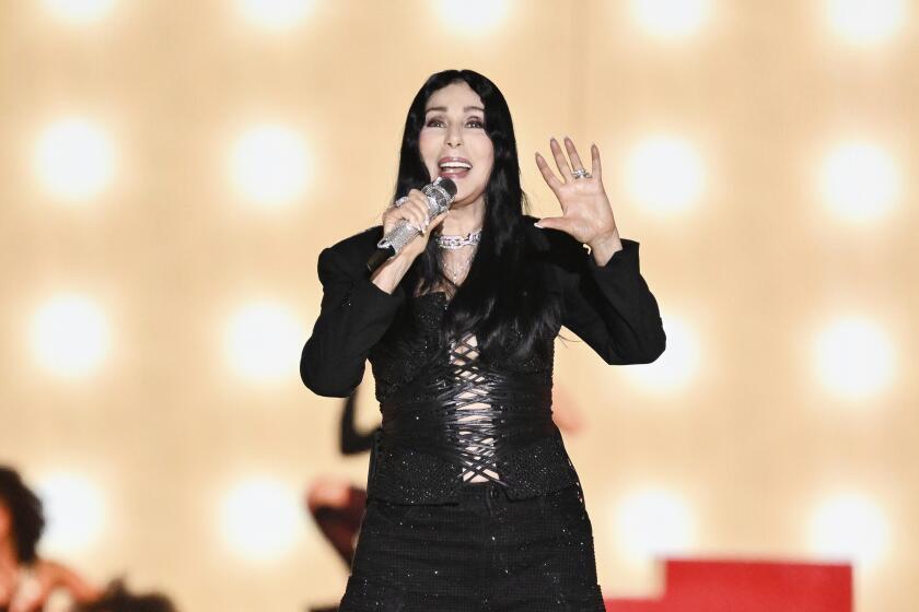 Cher sings into a microphone while holding up one hand in front of a lighted background