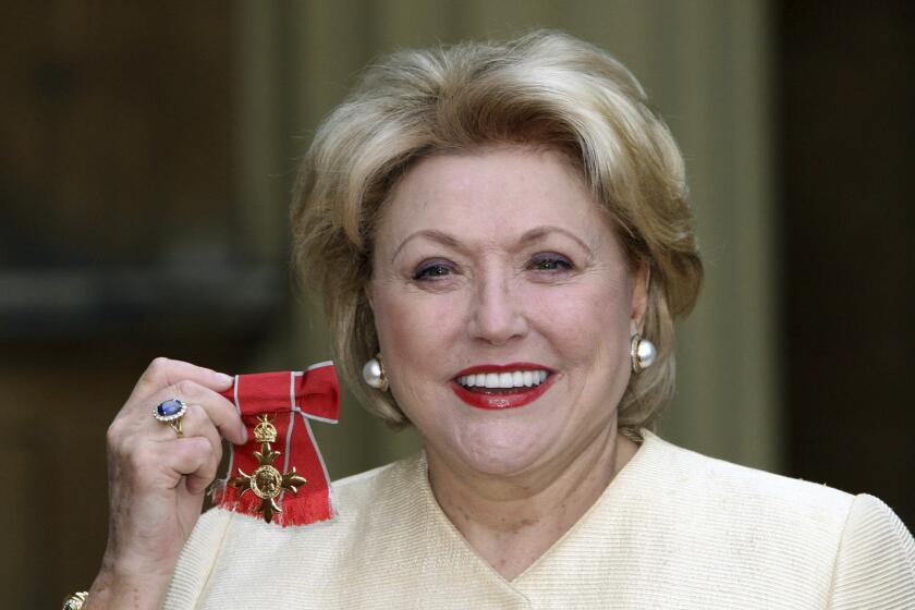 Barbara Taylor Bradford smiles in a cream suit while holding up a red British medal 